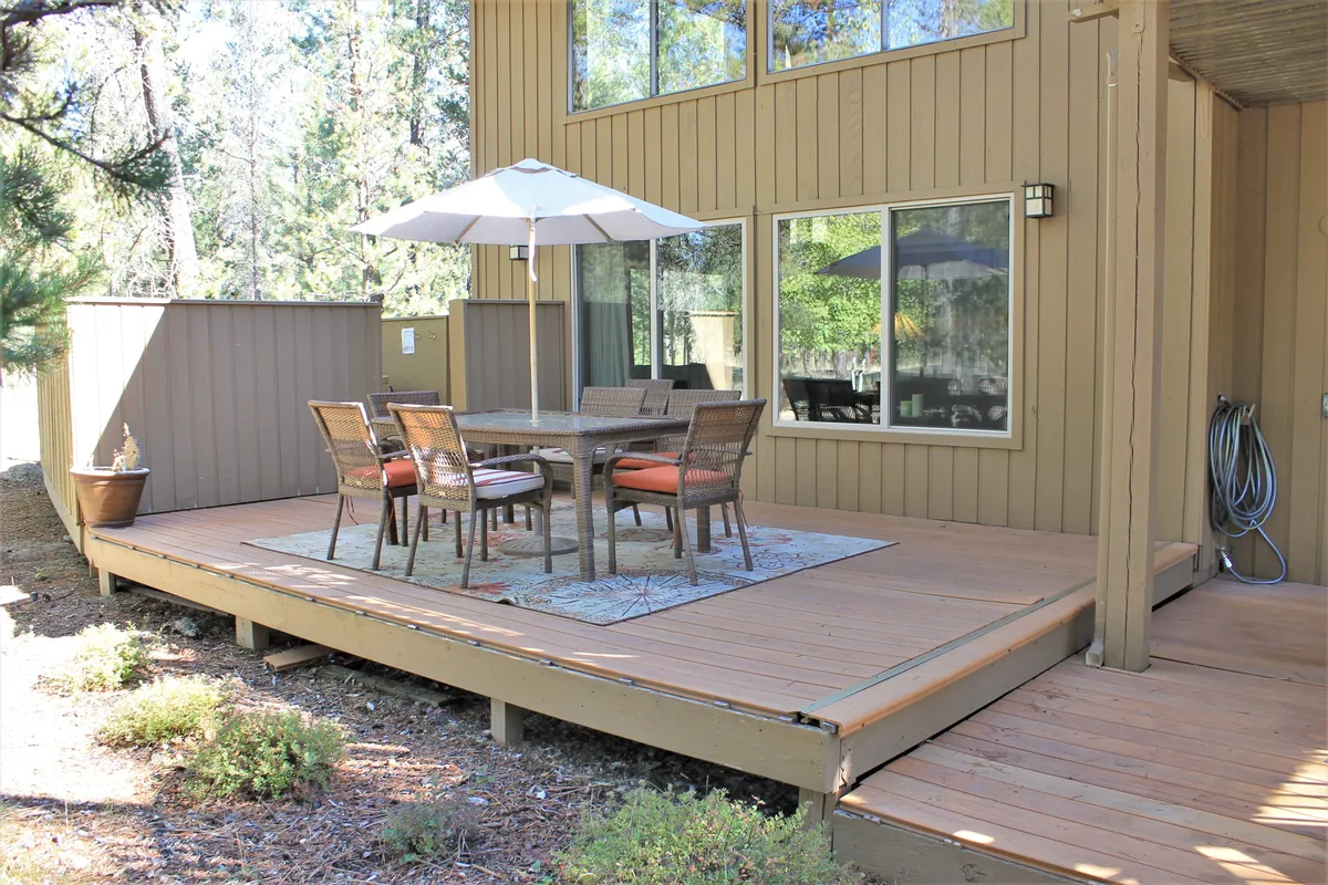 A spacious exterior deck surrounded by trees, featuring a large dining table shaded by an umbrella and ample seating, perfect for outdoor meals or gatherings. The scene exudes a relaxed and welcoming mood, ideal for socializing and enjoying nature.