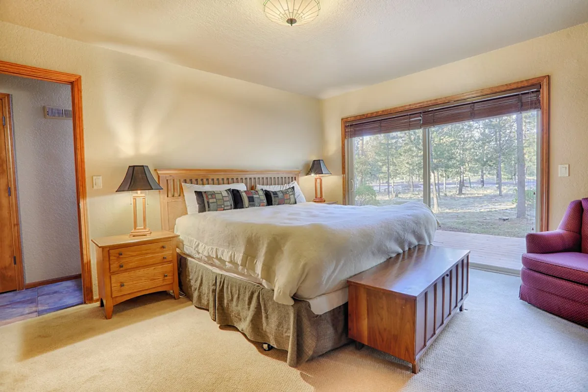 A cozy bedroom featuring a large bed with soft linens, nightstands with lamps, and an armchair positioned by a generous window, allowing natural light to flood the room. The tranquil décor and comfortable furnishings create an inviting, restful environment ideal for relaxation.