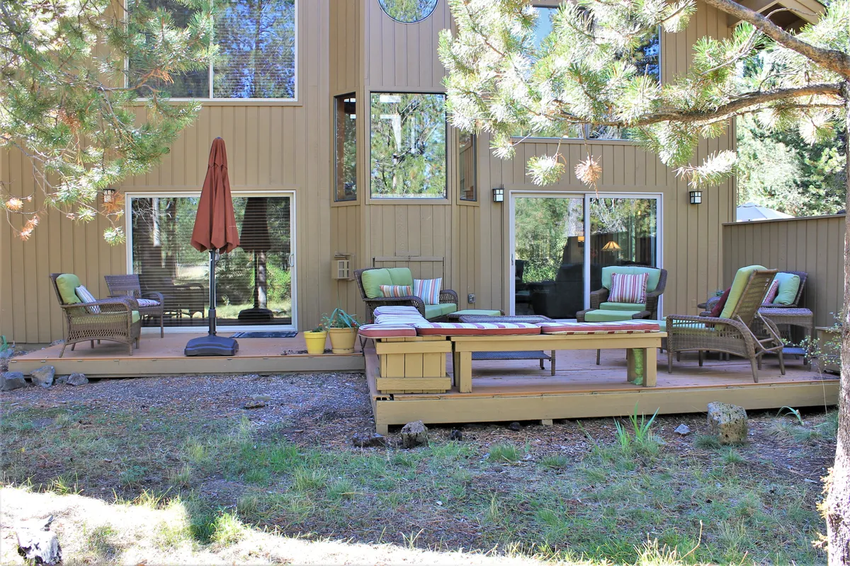 A tranquil patio seating area with comfortable outdoor furniture, an umbrella for shade, and views of the surrounding forest. The large deck and windows highlight the seamless connection between indoor and outdoor living.