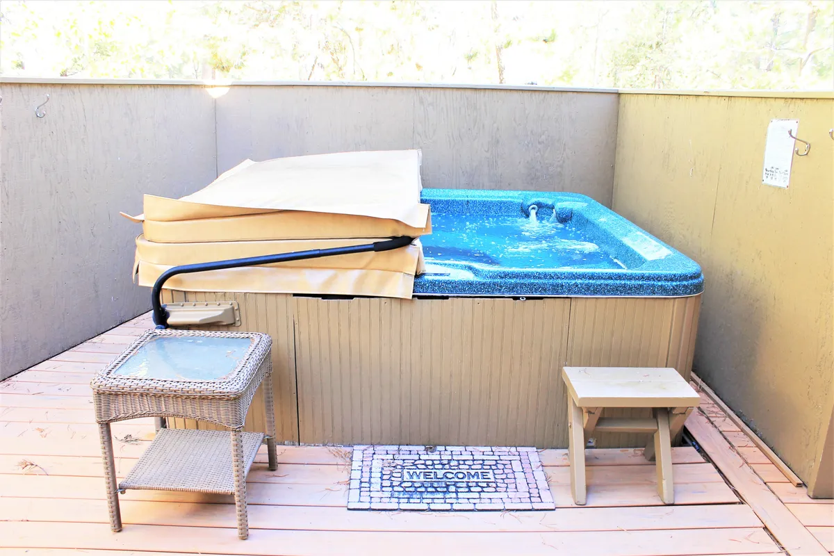 A cozy deck area featuring a private hot tub, welcome mat, small table, and surrounding wooden elements. The space is tranquil, inviting, and ideal for relaxing soaks after an active day, with potential for both group and solo enjoyment.