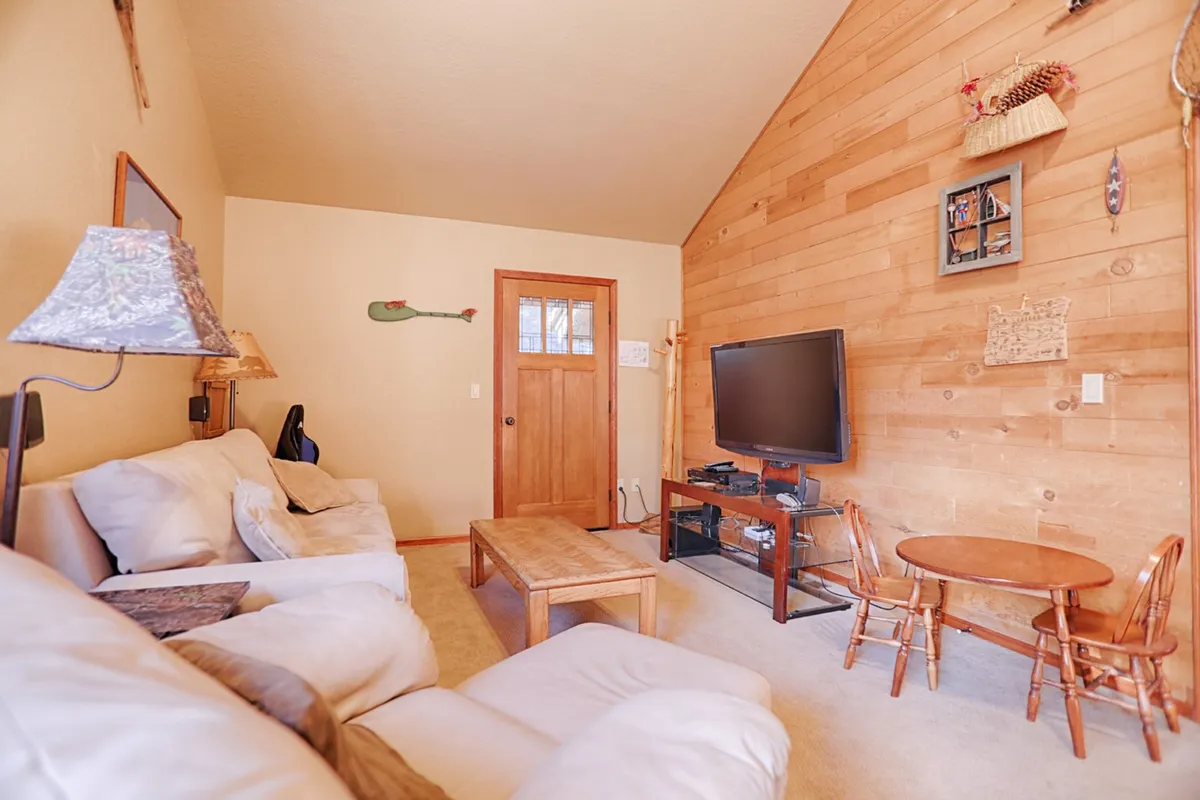 This image shows a comfortable seating area with a sofa, coffee table, TV, and dining table against a wooden accent wall. The setup suggests a place for both watching movies and playing games, with ample seating for gatherings.