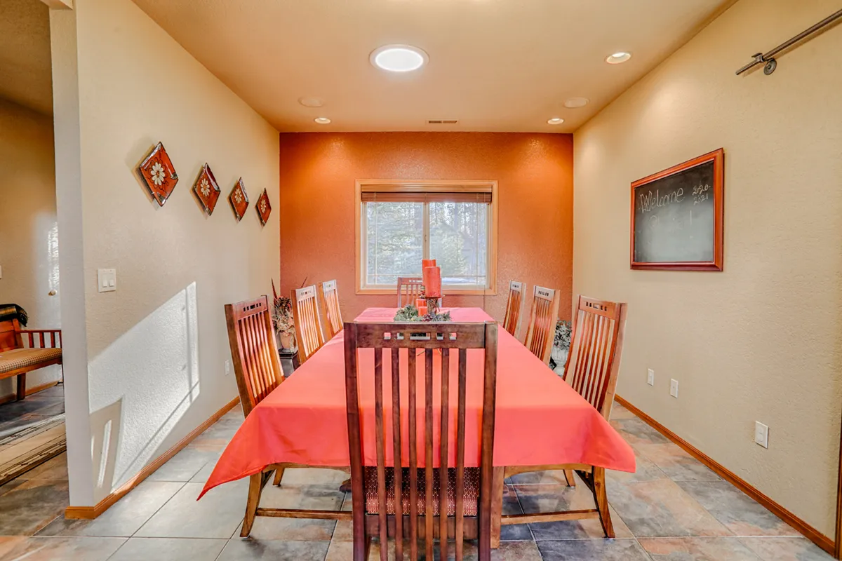 A long dining table draped with a red tablecloth is bathed in natural light from a nearby window. Decorative wall plates and a welcoming chalkboard add a touch of personality and warmth. The spacious setup and inviting decor clearly signal a space ready for large family meals and celebrations.