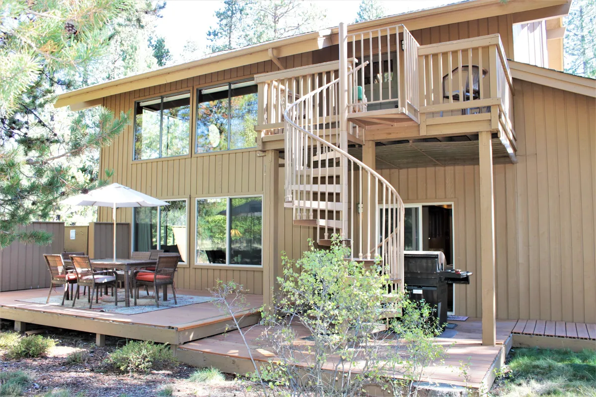 The upper deck features a spiral staircase, an outdoor dining set with a patio umbrella, and views of lush trees. The setup encourages outdoor meals, socializing, and enjoying the tranquil forested environment around the home.