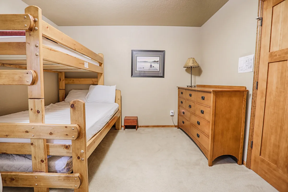 A cheerful bedroom with a sturdy bunk bed, a dresser, a playful framed picture, and a bedside lamp. The space feels inviting and is well-suited for children or family members seeking a cozy retreat of their own.