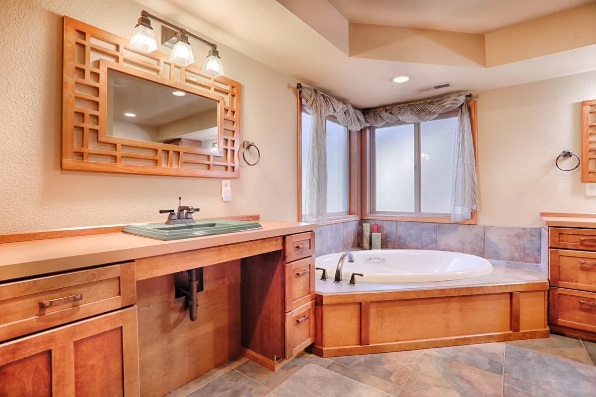 A master bathroom featuring a spacious tub, wooden cabinetry, a large mirror, and a washbasin, all bathed in soft natural light. The layout appears open and accessible, with thoughtful details that suggest a relaxing, inclusive environment.